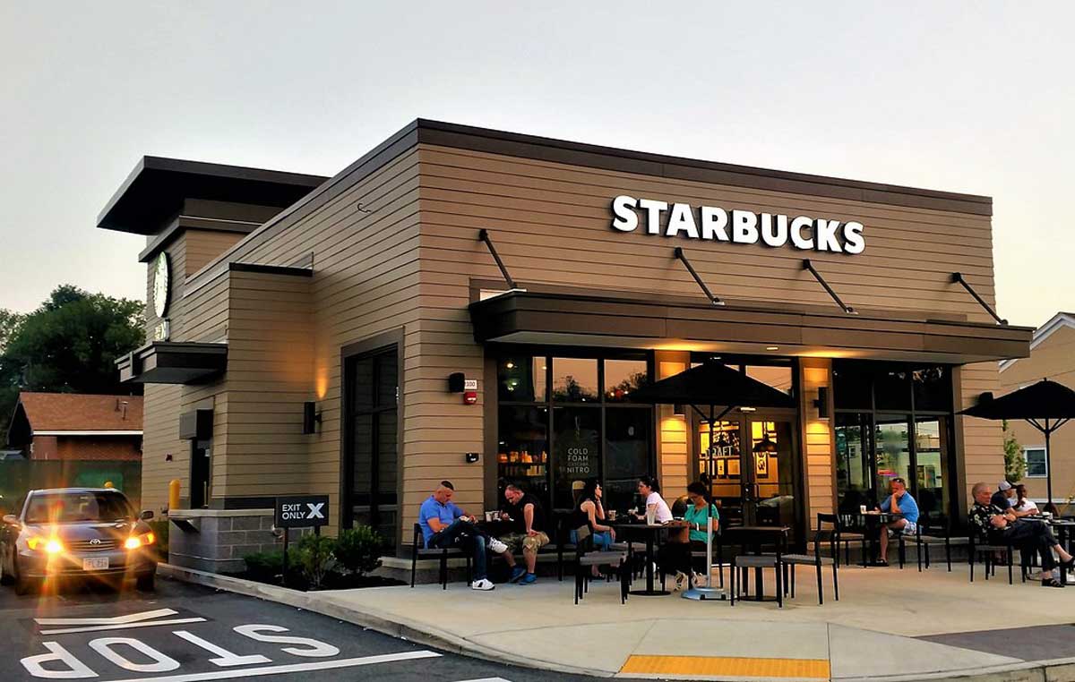 A modern Starbucks café with outdoor seating. Several people sit at tables under black umbrellas, enjoying drinks. A car waits near the exit, and evening light suggests it is near sunset.