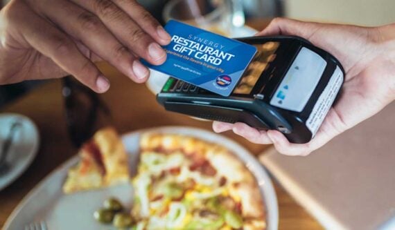 A person holds a restaurant gift card over a payment terminal, ready to pay. In the background, there is a plate of pizza on a wooden table.