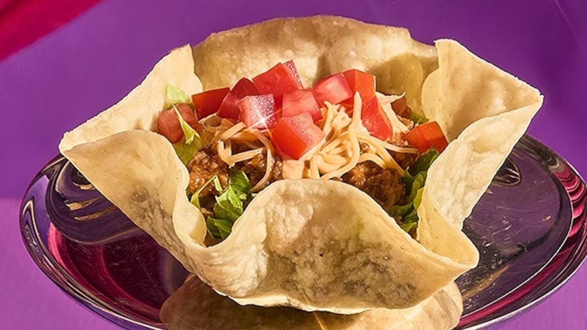 A crispy taco salad bowl filled with ground beef, shredded cheese, chopped tomatoes, and lettuce, served on a glass plate against a purple background.