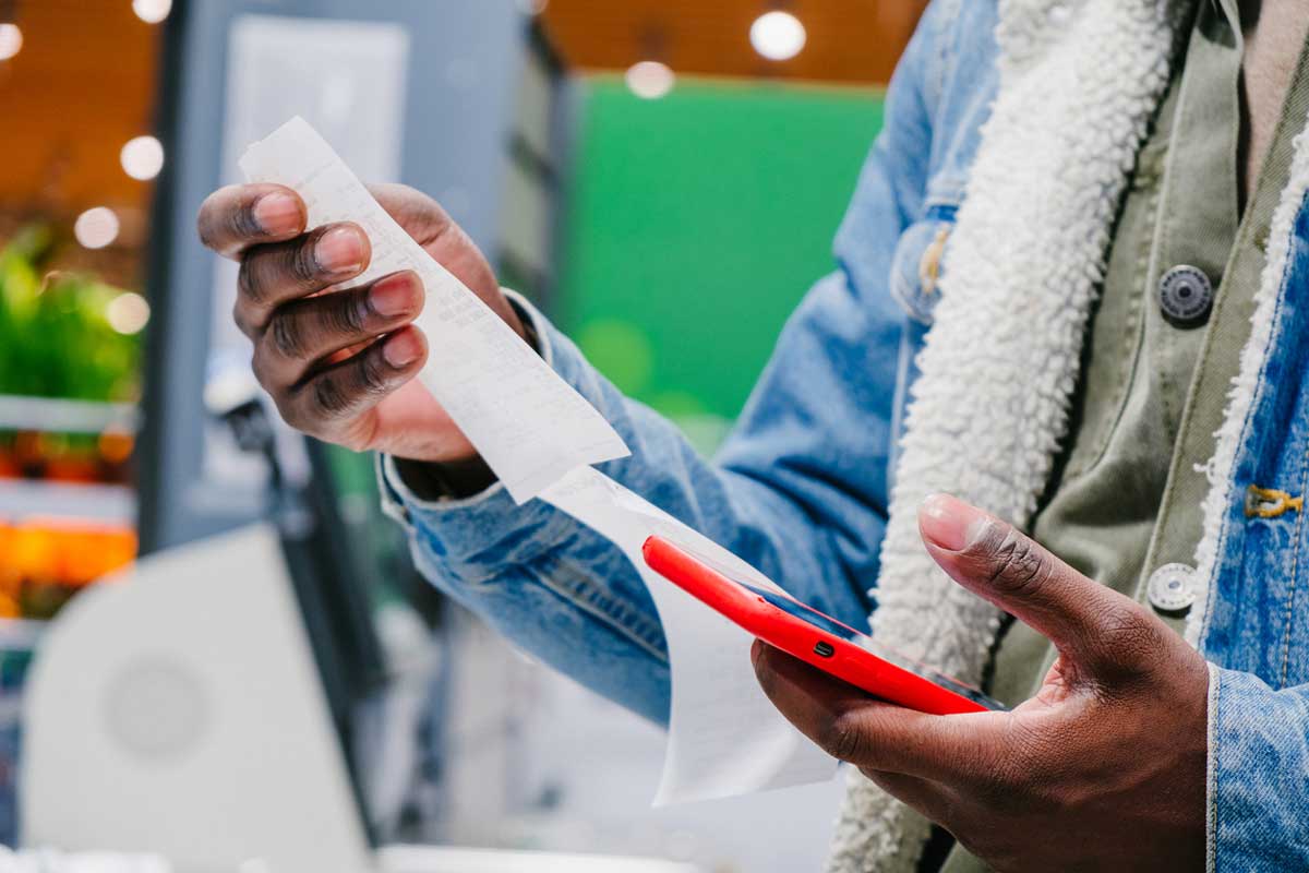 A person holding a red smartphone in one hand and examining a long receipt with the other hand, wearing a green shirt and a blue denim jacket with a shearling lining.