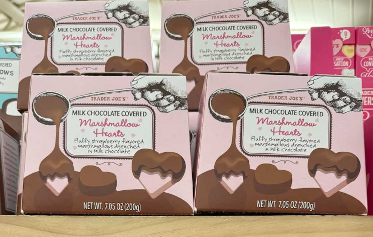 Boxes of Trader Joe’s Milk Chocolate Covered Marshmallow Hearts are stacked on a shelf. The pink packaging features heart-shaped chocolates and handwritten-style text describing the product.