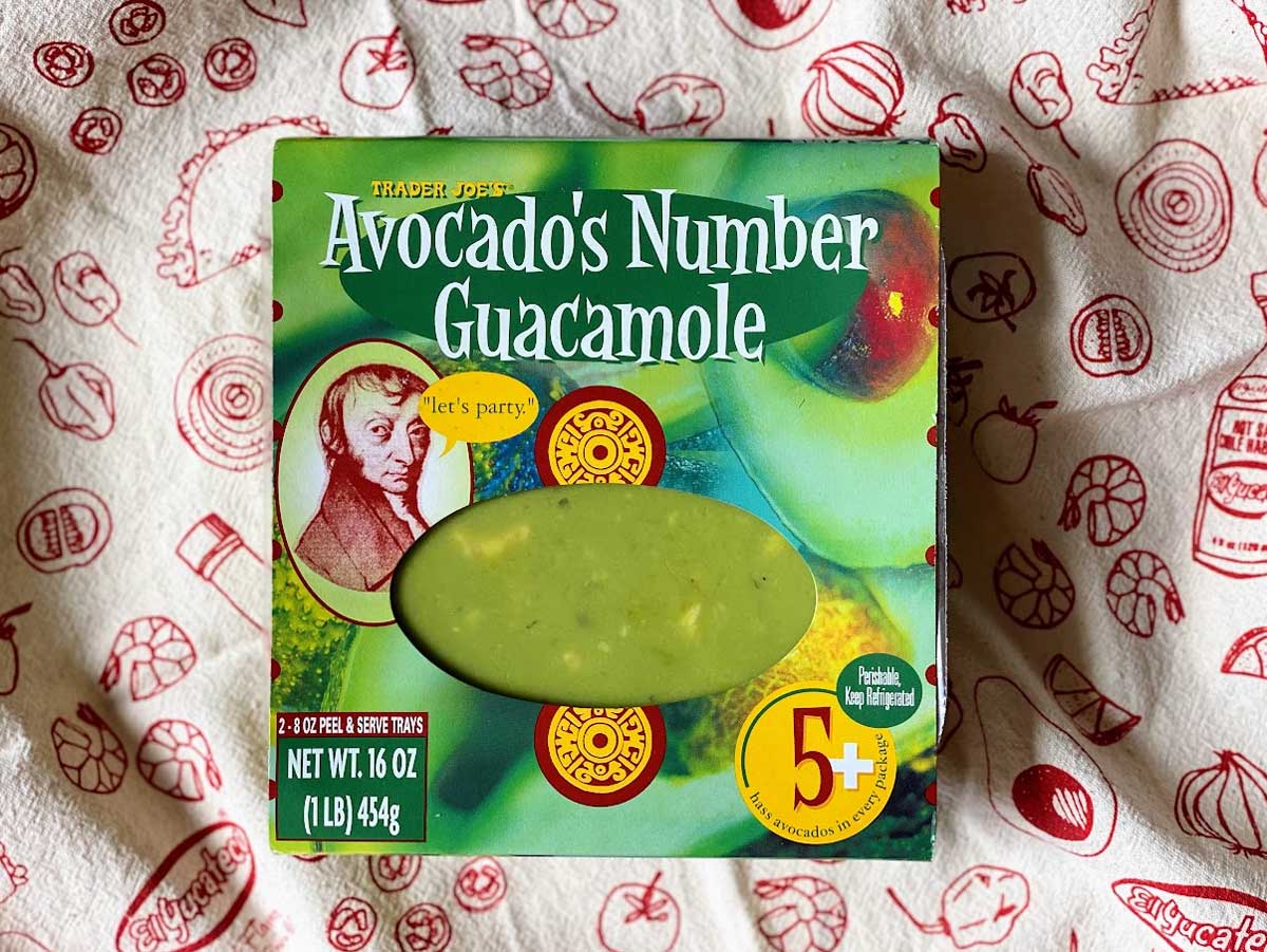 A box of Trader Joe’s Avocado’s Number Guacamole is placed on a patterned tablecloth. The packaging is green with an avocado graphic, nutrition facts, and a clear window showing the guacamole inside.