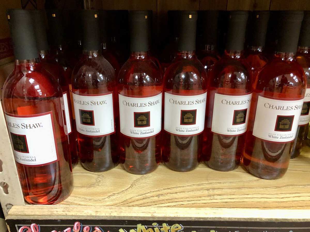 Several bottles of Charles Shaw White Zinfandel wine are lined up on a wooden shelf, with their labels clearly visible and the pink rosé liquid inside the bottles.