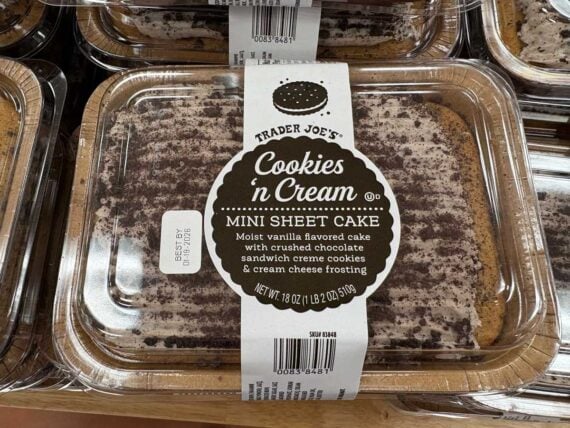 A clear plastic container holds a Trader Joe's Cookies n Cream Mini Sheet Cake, topped with crushed chocolate sandwich cookies and cream cheese frosting. A white label with product details is centered on top.