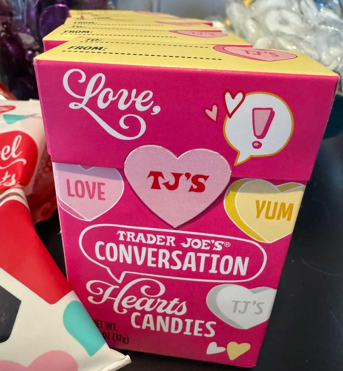 A pink box of Trader Joe's Conversation Hearts Candies decorated with colorful heart shapes and words like "LOVE," "YUM," and "TJ'S" on the front. The box is displayed on a store shelf.