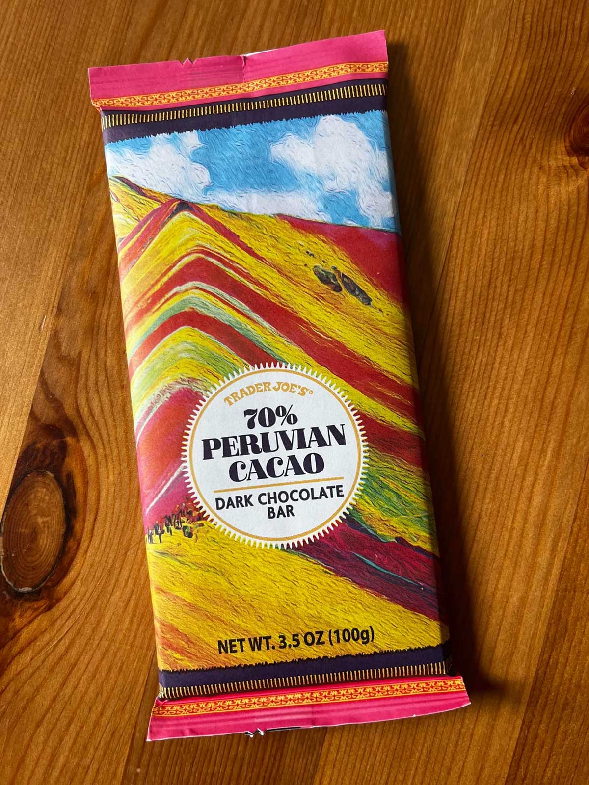 A Trader Joe’s 70% Peruvian Cacao dark chocolate bar lies on a wooden surface. The colorful wrapper features an illustration of Peru’s Rainbow Mountain and a circular label with product details.