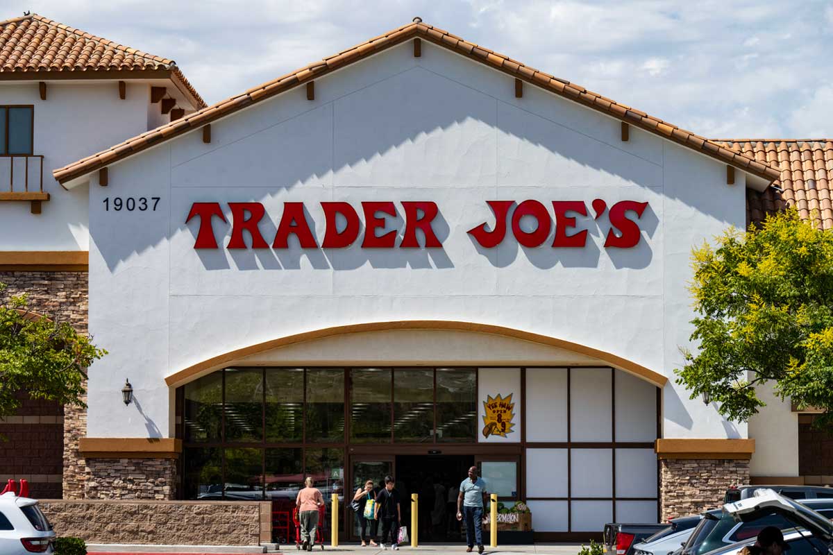 The front of a Trader Joe’s store with large red signage and several people entering and exiting the entrance under a partly cloudy sky. Cars are parked in front of the building.