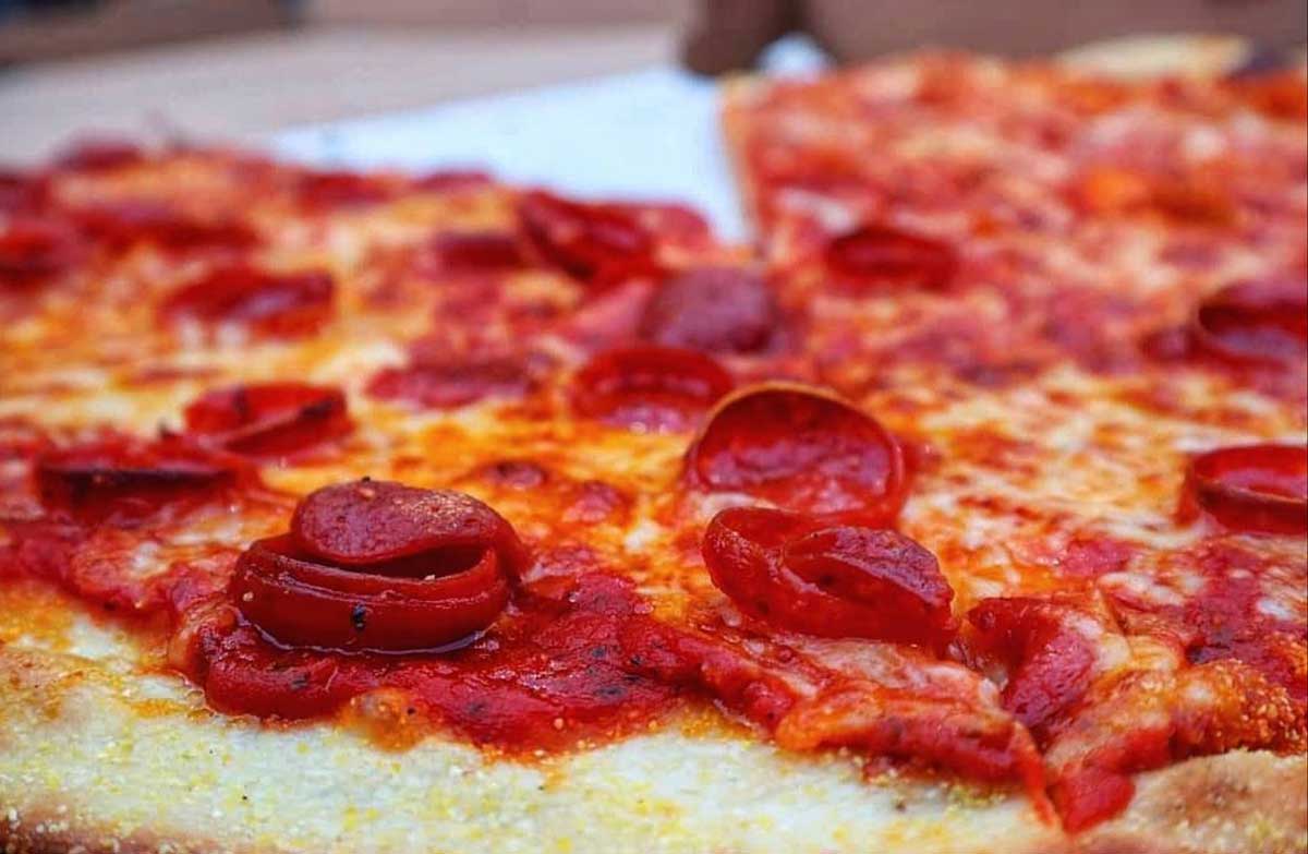 Close-up of an east coast cheap pizza slice, showing melted cheese, crispy pepperoni, and a golden, slightly thick crust. The pizza looks fresh and inviting, with a rich layer of tomato sauce.