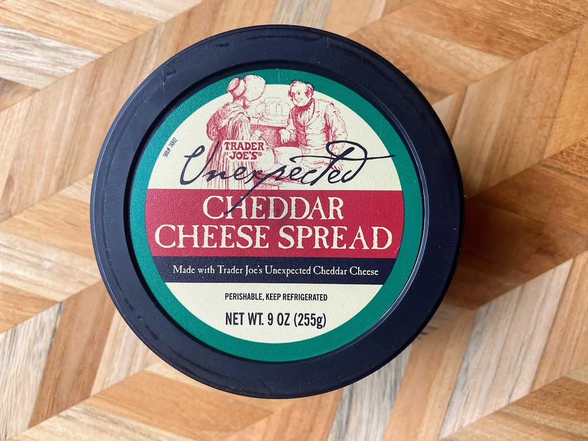 A round container of Trader Joe's Unexpected Cheddar Cheese Spread sits on a wooden, geometric-patterned surface. The label is green and red with vintage-style illustrations and product details.