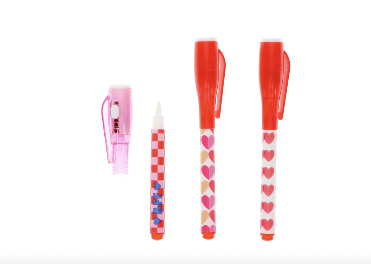 Three colorful pens with red caps and heart or checkered designs stand upright on a white background, next to a detached pink pen cap lying on its side.