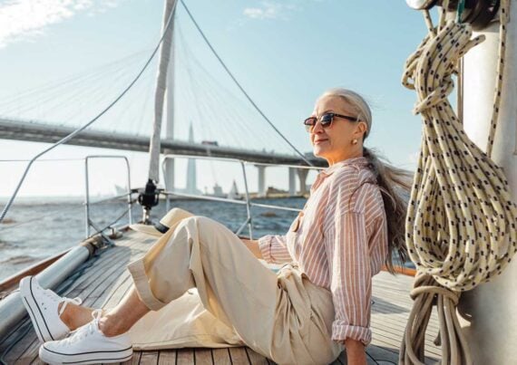 An older woman with long gray hair, sunglasses, and casual clothes relaxes on a sailboat deck. A bridge and city skyline appear in the background on a sunny day.