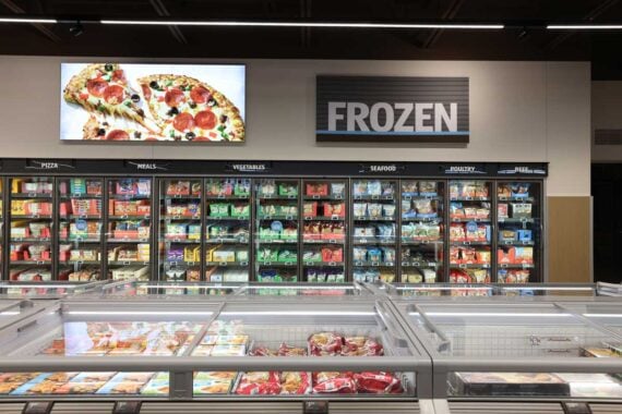A supermarket frozen food aisle with glass-door freezers stocked with various frozen foods. A large sign reads "FROZEN," and a digital display above shows an image of a pizza. Frozen products are organized by category.