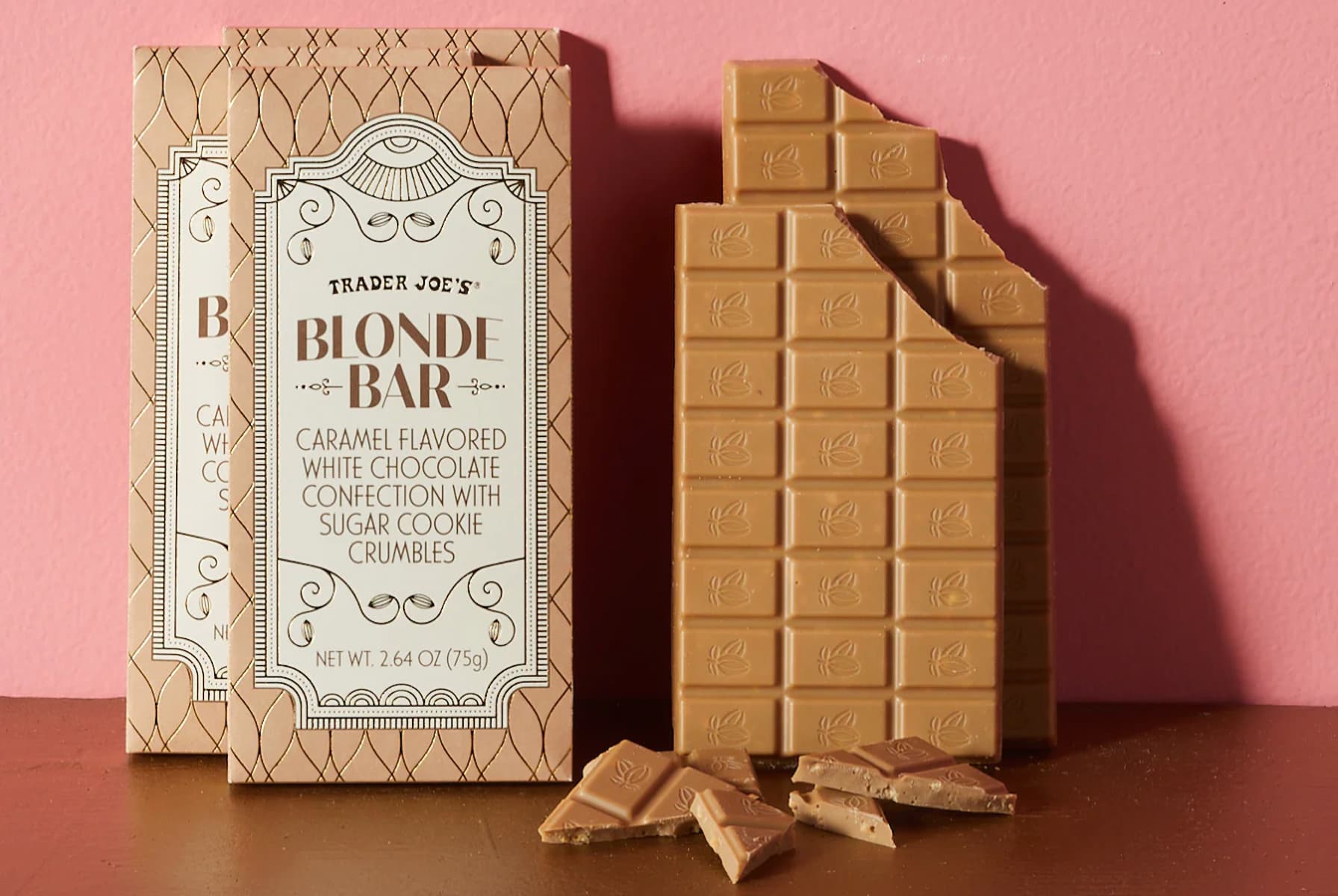 A Trader Joe's Blonde Bar package stands next to a partially broken caramel-colored chocolate bar with several pieces scattered on a brown surface, set against a pink background.