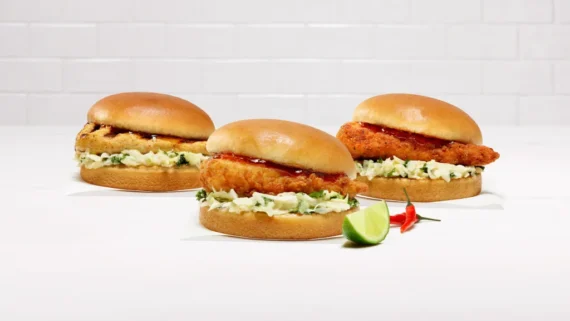 Three fried chicken sandwiches with lettuce and sauce on buns are arranged on a white surface, with a sliced lime and two red chili peppers placed beside them. The background is a white tiled wall.