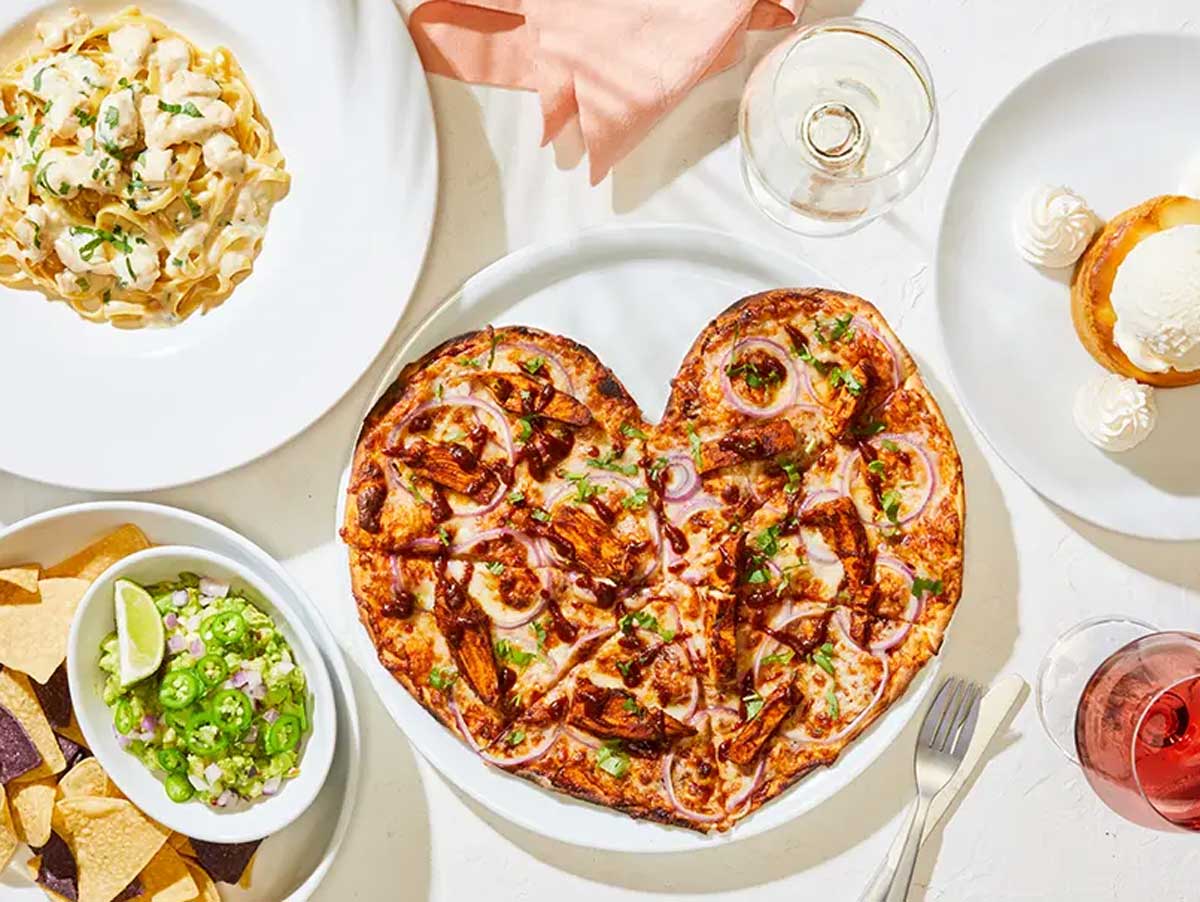 A heart-shaped pizza with onions and green peppers is surrounded by a plate of pasta, chips with dip, a dessert with ice cream, and glasses of wine on a white table.