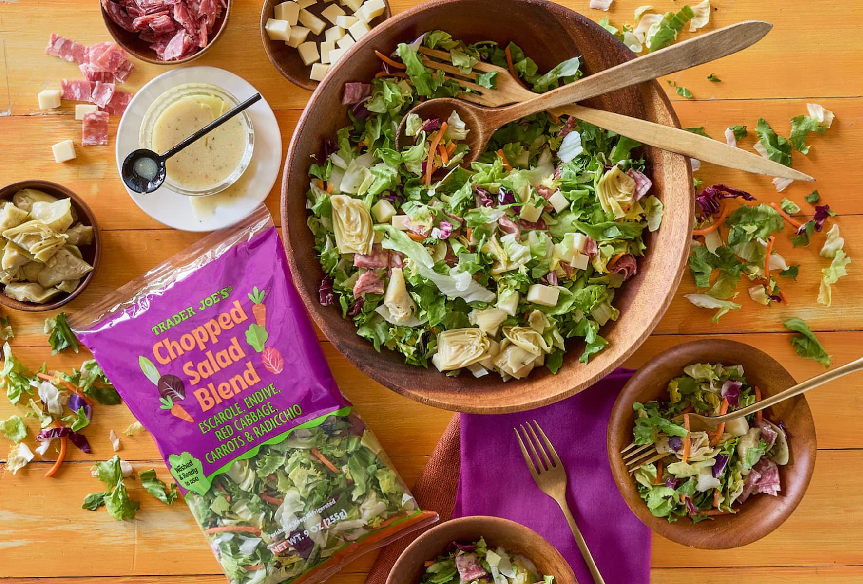 A wooden bowl of chopped salad with artichokes, cheese cubes, and salami sits on a table with wooden salad servers, a smaller salad bowl, dressing, and an open Trader Joe’s Chopped Salad Blend bag nearby.