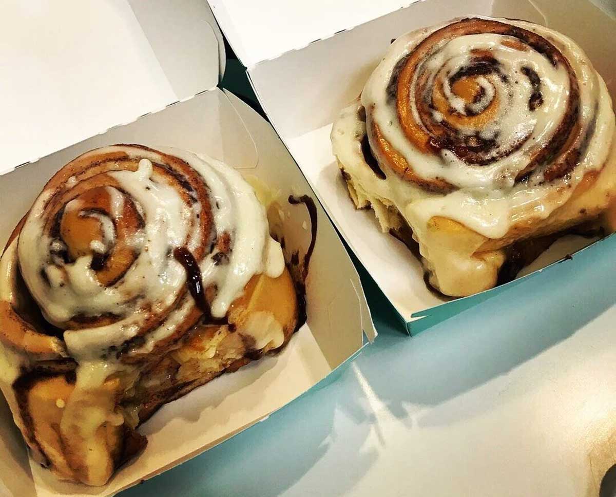 Two cinnamon rolls topped with creamy white icing sit in individual open boxes on a light-colored surface. The rolls appear soft, gooey, and freshly baked, with swirls of cinnamon visible.
