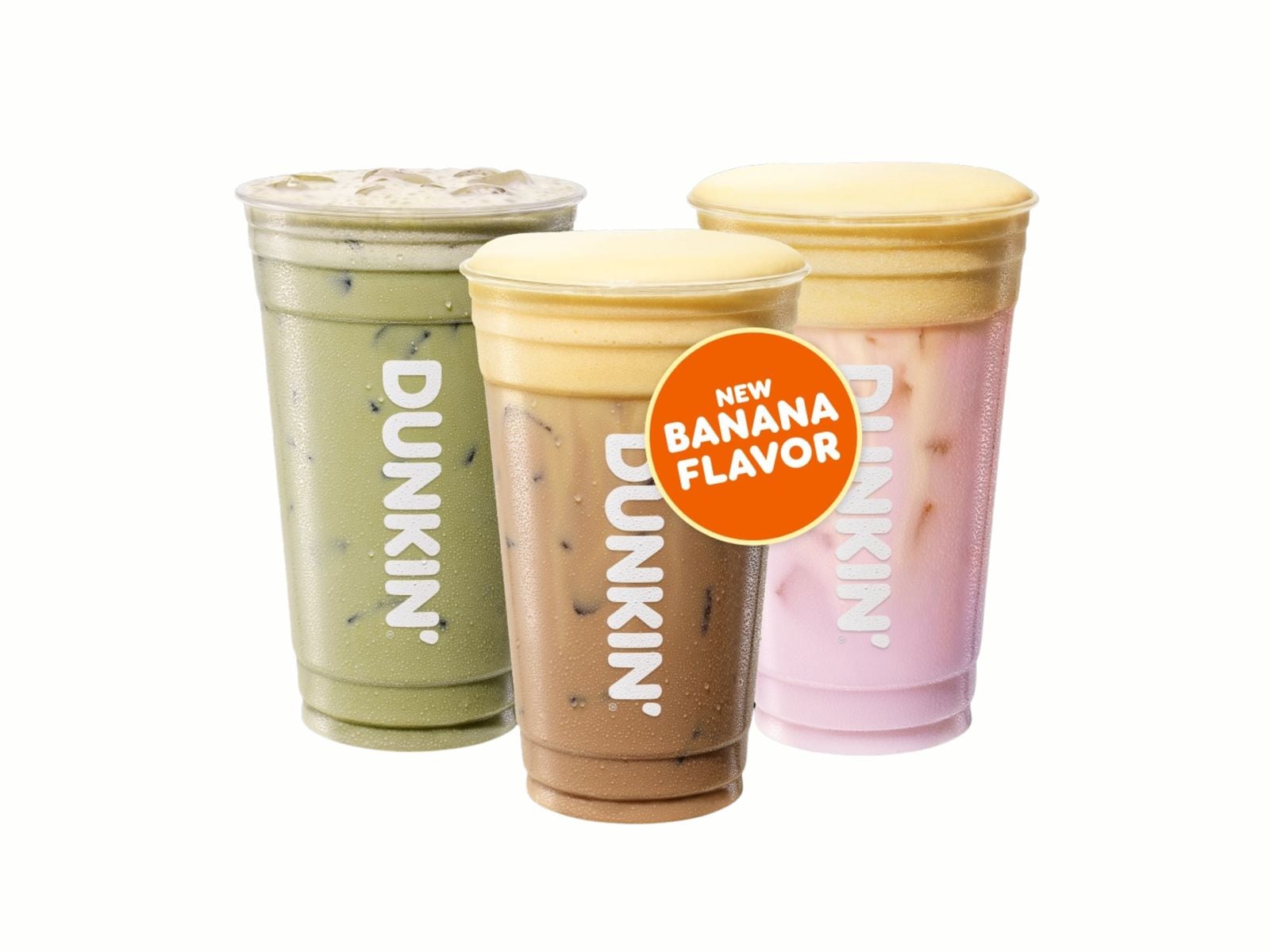 Three Dunkin’ iced drinks in green, brown, and pink with the Dunkin’ logo on each cup. An orange sticker on the pink drink says "NEW BANANA FLAVOR," highlighting a tasty addition to the new fast-food menu. The background is white.