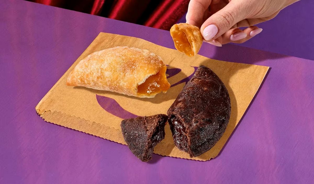 A hand holds a piece of a golden-brown empanada with filling, next to a whole empanada and a halved dark chocolate empanada—all part of an exciting new fast-food menu—served on a brown paper bag atop a purple surface.