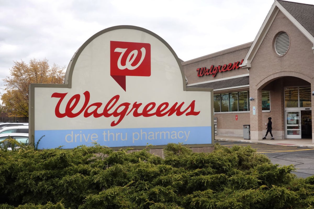 A large Walgreens sign reading "drive thru pharmacy" stands in front of the brick store. Amid news of 2026 layoffs, shrubs line the foreground and a person enters the store under cloudy skies.