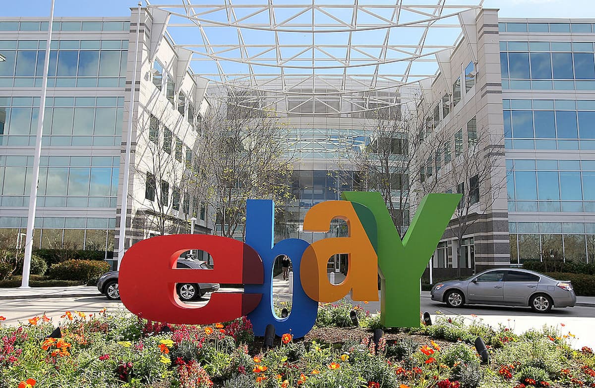 The eBay headquarters building, with a large, colorful eBay logo sculpture in front and cars parked nearby, stands amid flowers. Its glass windows reflect the blue sky—a site witnessing changes like the 2026 layoffs.