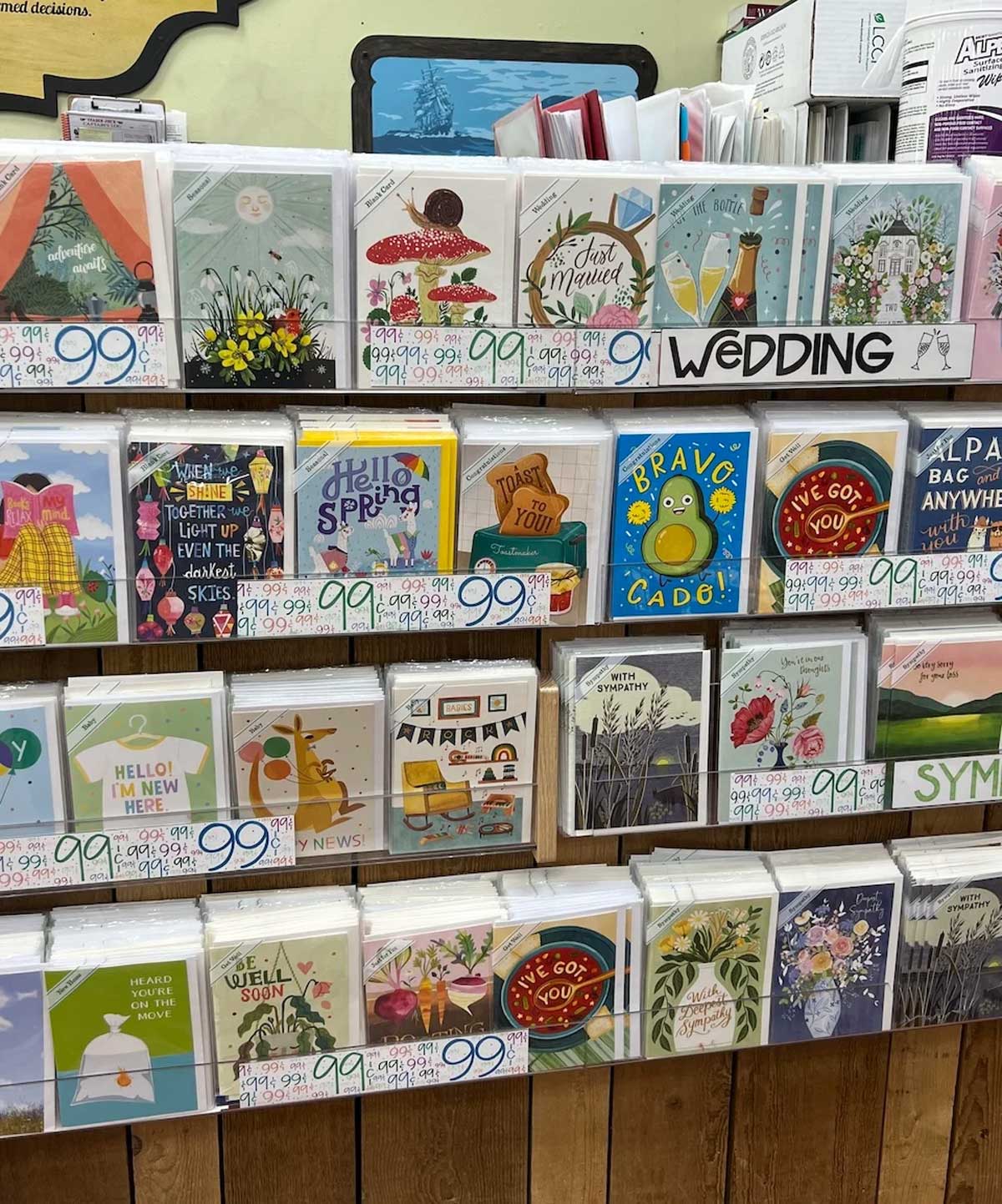 A display of colorful greeting cards with various designs and messages, including wedding, spring, congratulations, and encouragement themes. Each card is priced at 99 cents and organized neatly on wooden shelves.