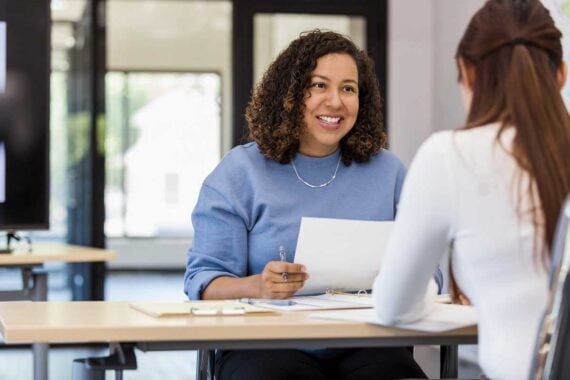 A woman with curly hair, wearing a blue sweater, smiles while holding papers and talking to another woman with long brown hair across a desk in a bright office setting.