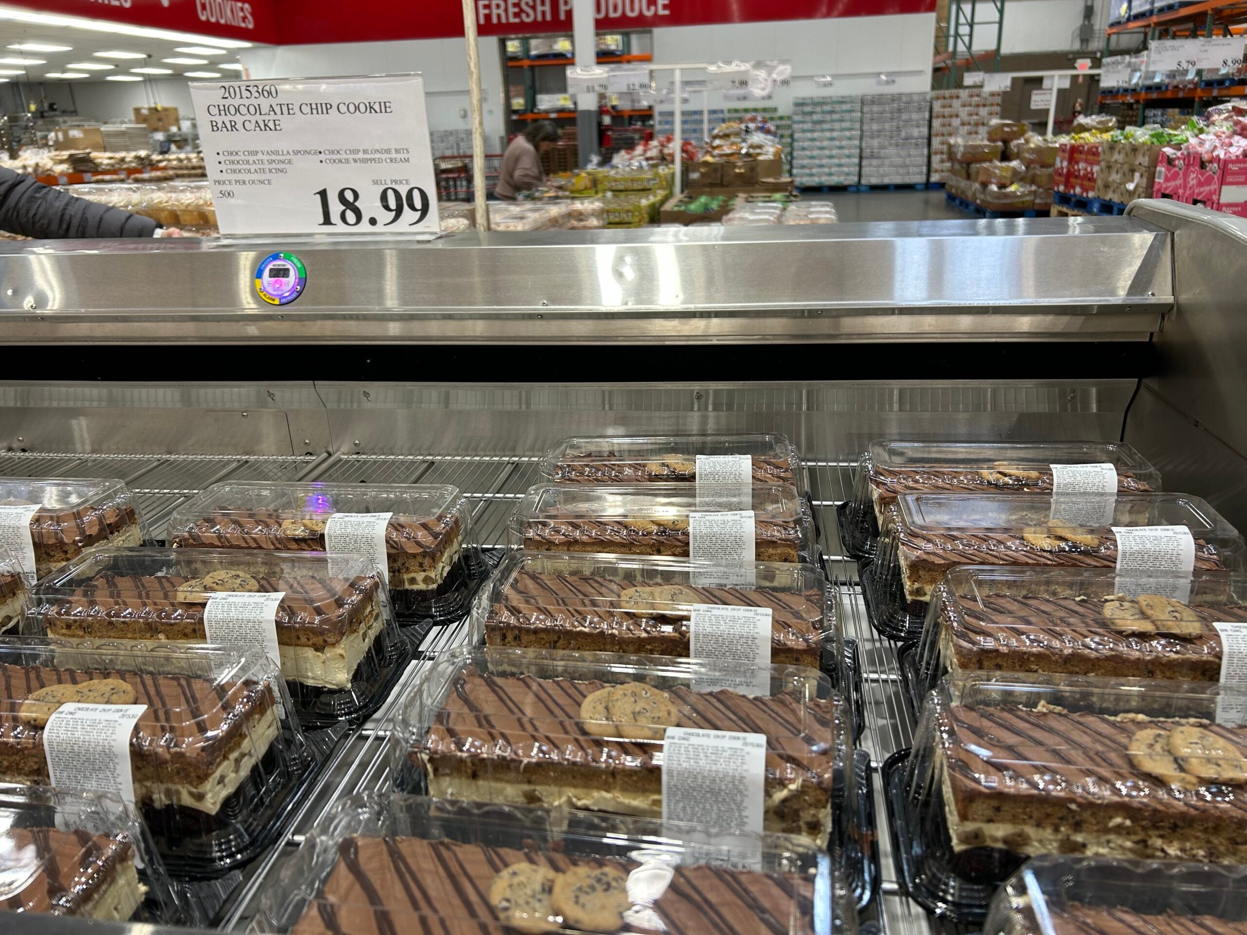 Chocolate chip cookie bar cakes in clear plastic containers are displayed on a refrigerated shelf at a store. A sign above reads "Chocolate Chip Cookie Bar Cake $18.99." Other store products are visible in the background.