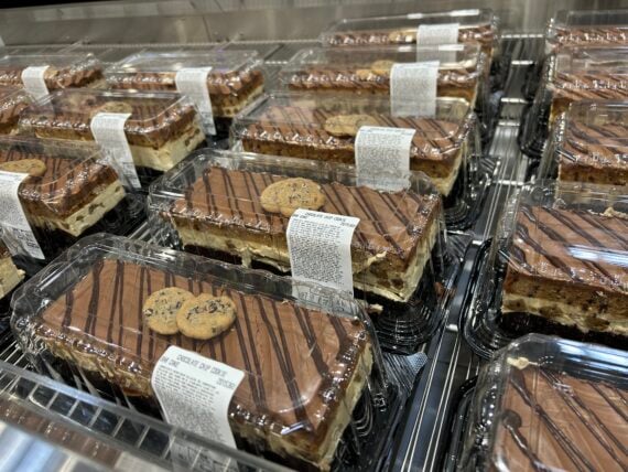 Rectangular chocolate cakes in clear plastic containers are displayed on metal shelves. Each cake is topped with two chocolate chip cookies and drizzled with chocolate. White labels with text are on top of the containers.
