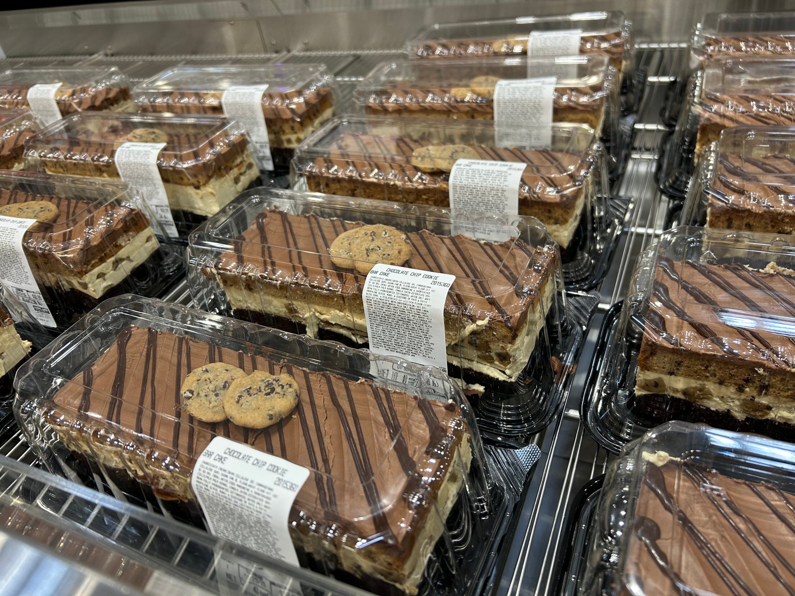 Rectangular chocolate cakes in clear plastic containers are displayed on metal shelves. Each cake is topped with two chocolate chip cookies and drizzled with chocolate. White labels with text are on top of the containers.