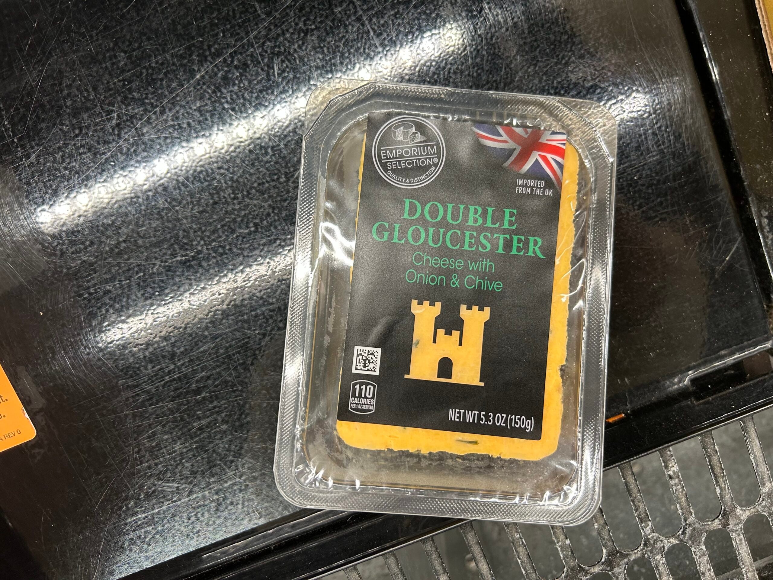 A package of Emporium Selection Double Gloucester cheese with onion and chive, weighing 5.3 oz (150g), sits on a black surface. The packaging features a castle icon and a Union Jack flag.