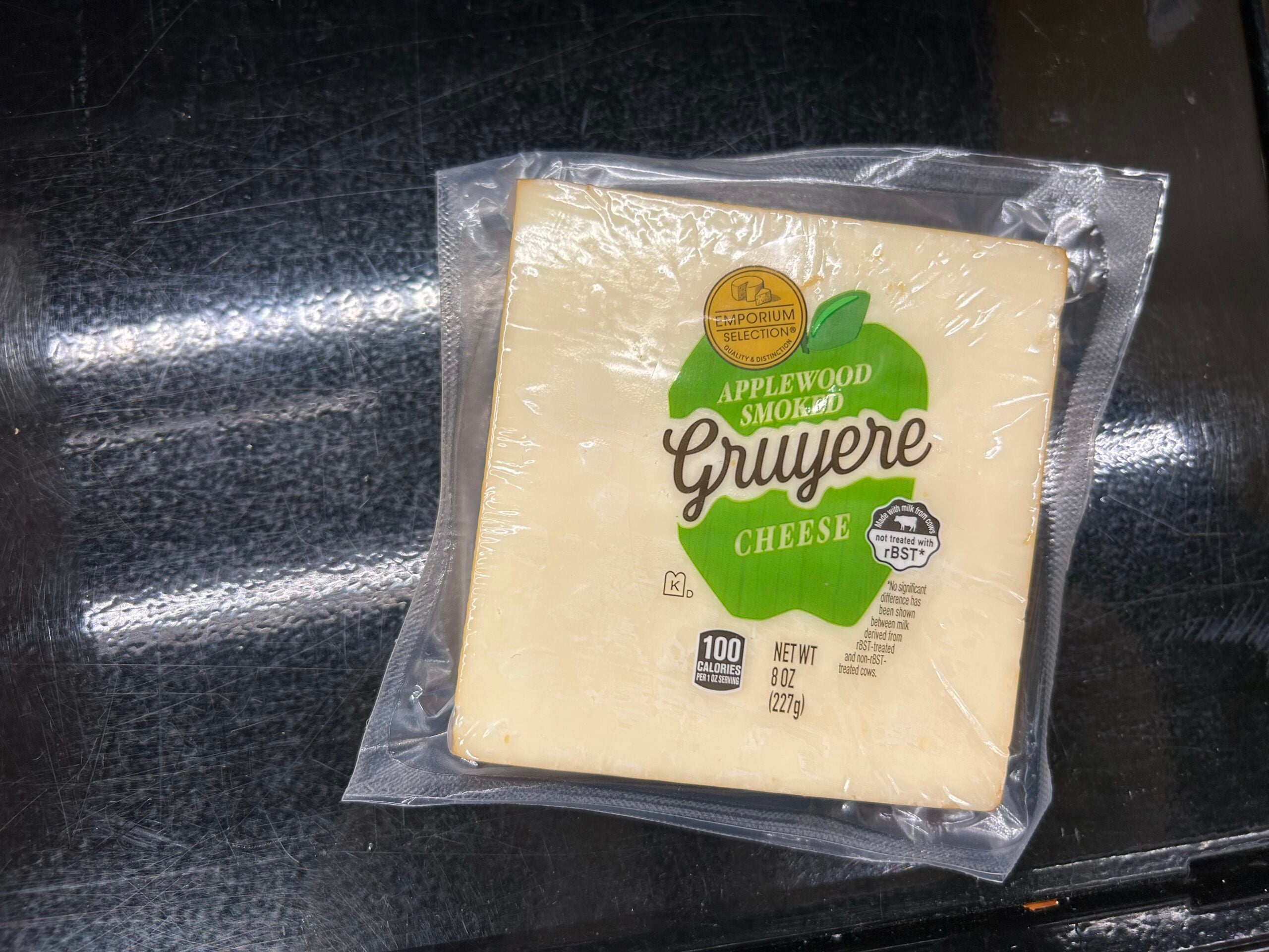 A sealed package of Applewood Smoked Gruyere cheese sits on a dark surface. The label shows the cheese weighs 8 oz (227g) and contains 100 calories per serving.