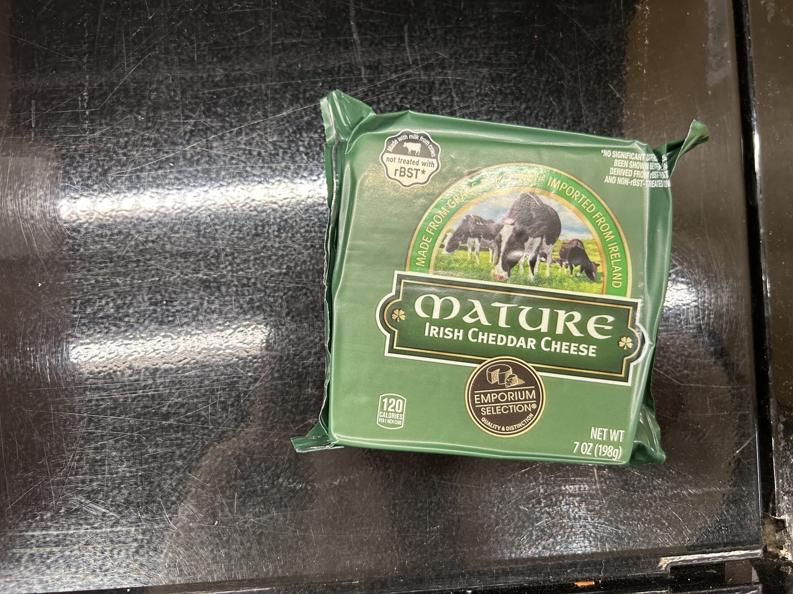 A green package of Mature Irish Cheddar Cheese from Emporium Selection sits on a black surface. The label features cows grazing in a field and states the cheese is imported from Ireland. The package weighs 7 oz (198g).