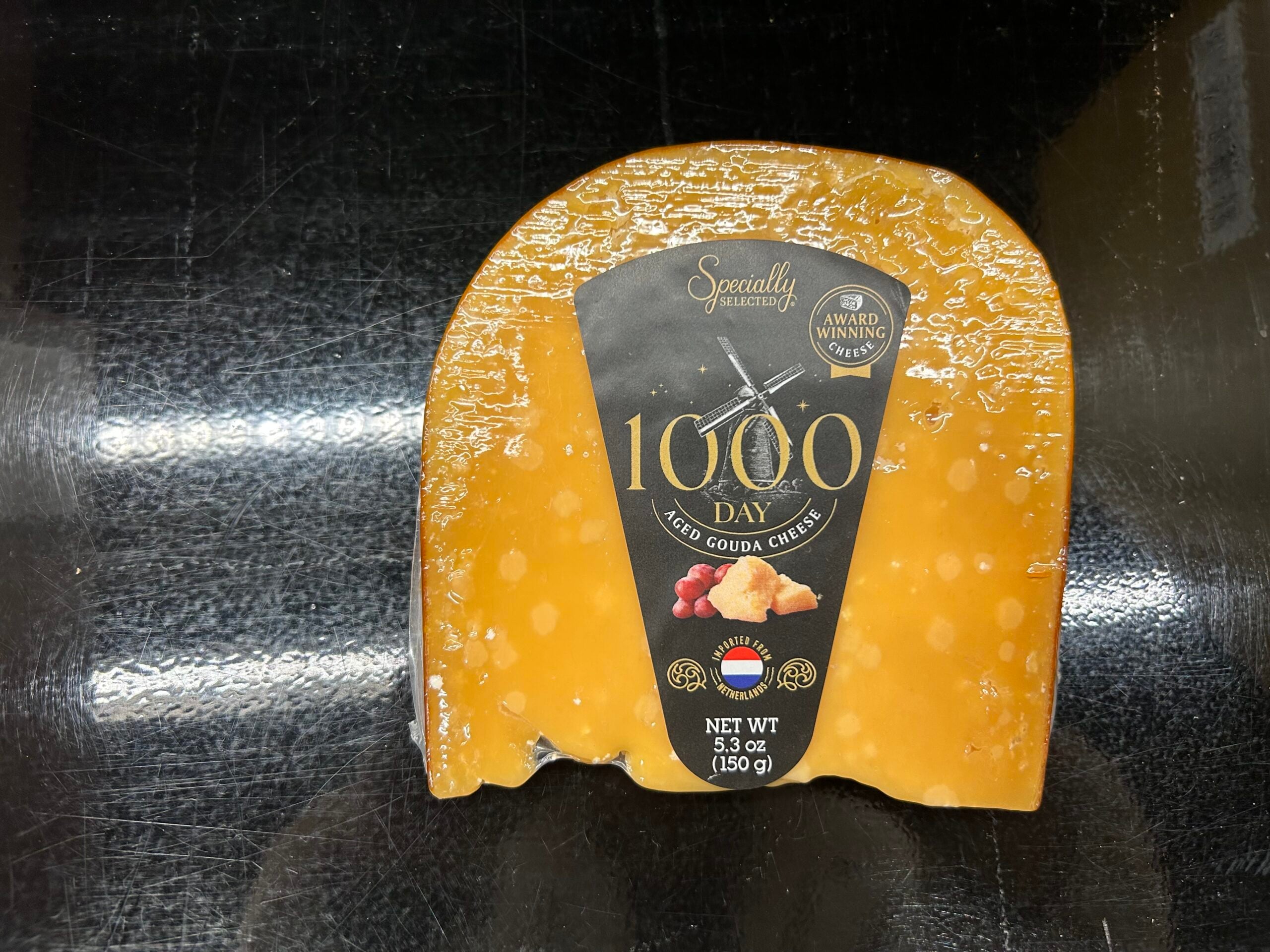 A wedge of 1000 Day aged gouda cheese with a black label reading “Specially Selected,” “Award Winning,” and weighing 5.3 oz (150 g), placed on a black reflective surface.
