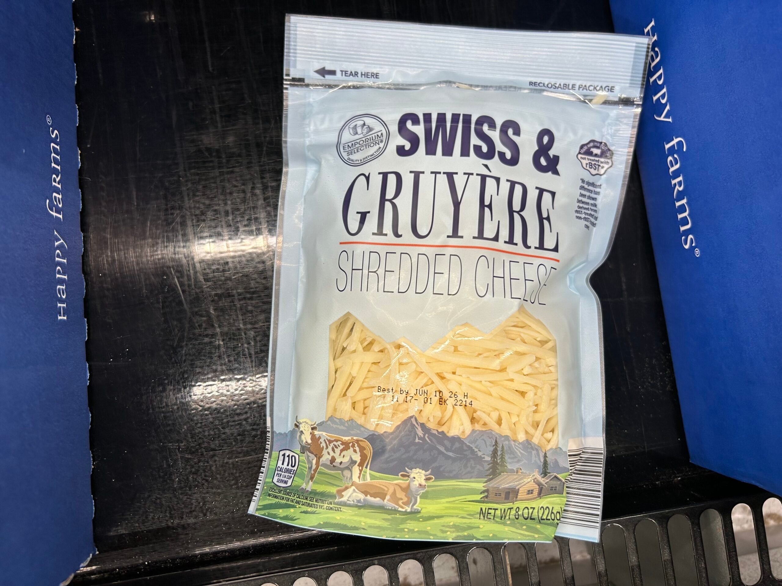 A bag of Happy Farms Swiss & Gruyere shredded cheese sits on a checkout conveyor belt. The packaging shows shredded cheese and an illustration of cows grazing in front of mountains.
