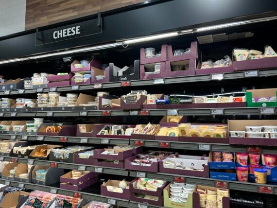 A supermarket refrigerated section filled with various cheeses and dairy products in different shapes and packaging, all neatly arranged on shelves beneath a sign labeled "CHEESE.