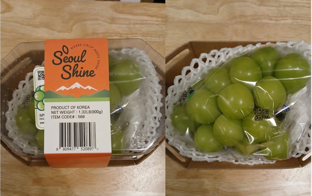 A plastic container of green grapes labeled “Seoul Shine” with orange and white branding. The grapes are packed in a tray with protective white padding and wrapped in clear plastic. The label shows weight and product info.