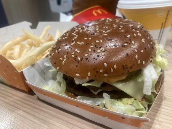 A close-up of a McDonald's meal featuring a sesame seed burger with lettuce, cheese, and sauce in a box, served with French fries and a drink in the background on a wooden table.