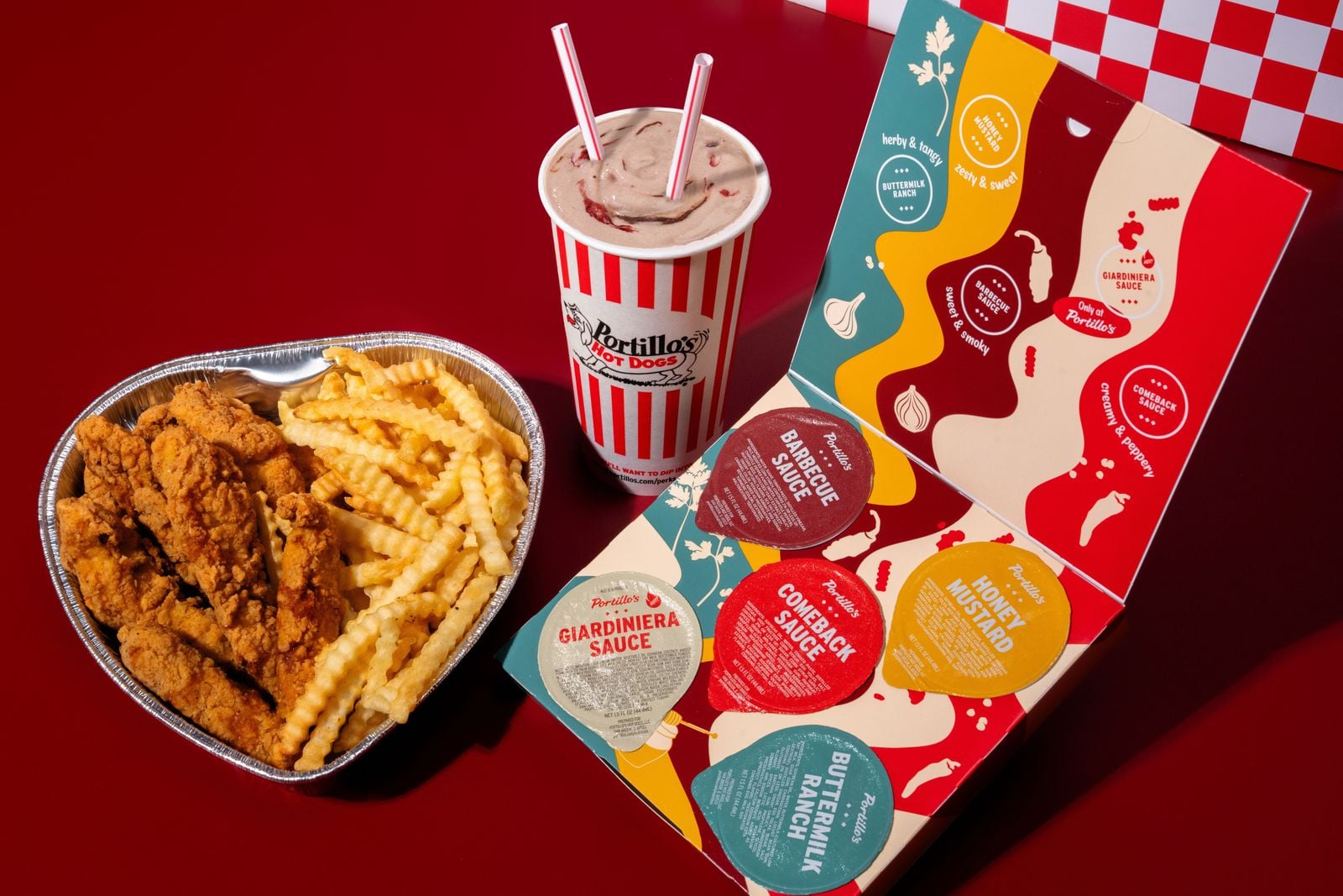 A heart-shaped tray holds crispy chicken tenders and crinkle-cut fries. Next to it is a large milkshake with two straws and a colorful open box displaying four dipping sauces from Portillo’s, all set on a red surface.