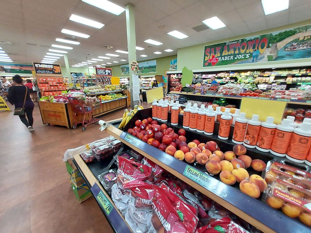 A grocery store produce section displays peaches, nectarines, and bottles of juice or smoothie. Colorful fruits and vegetables fill the shelves; a shopper walks down the aisle. A mural above reads "San Antonio Trader Joe's.