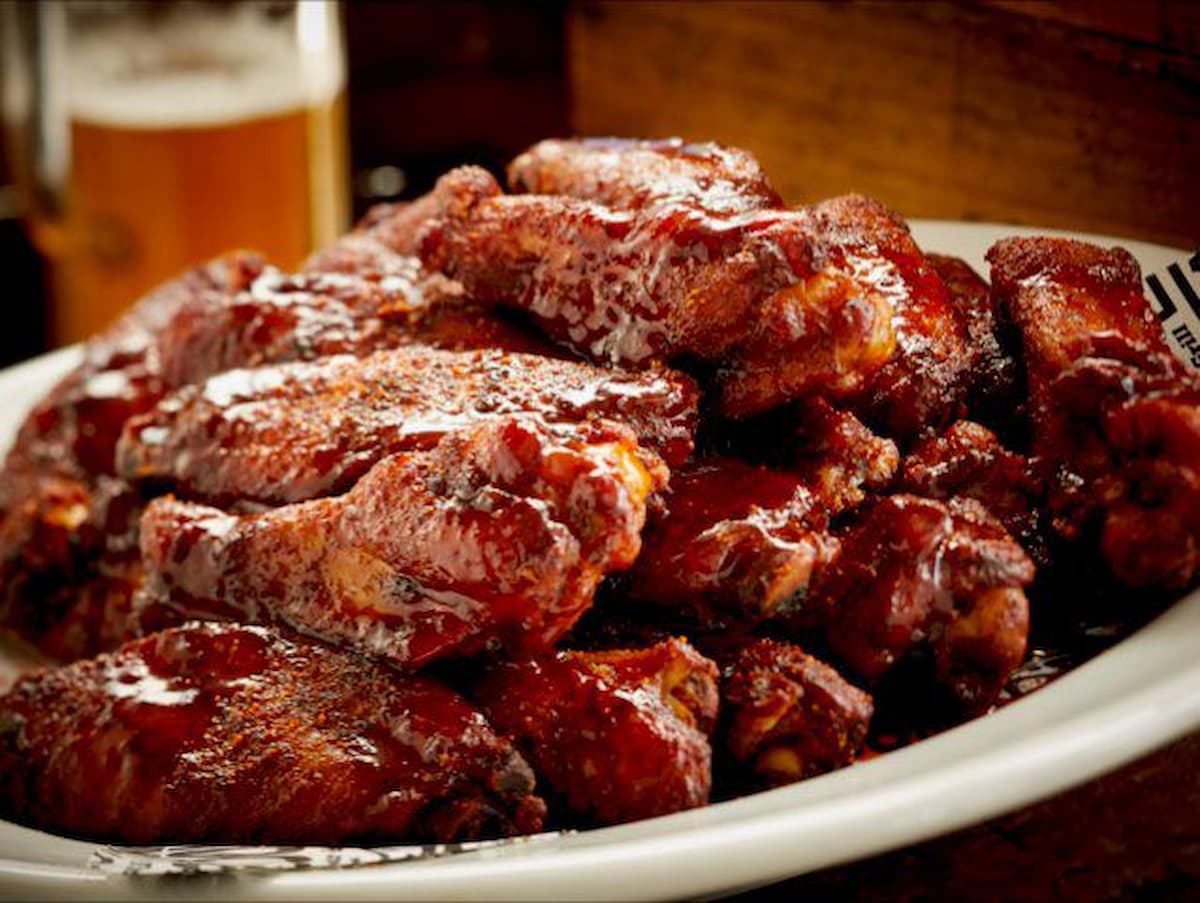 A plate piled high with glazed, barbecue chicken wings, glistening with sauce. A glass of beer is blurred in the background, suggesting a casual dining setting.