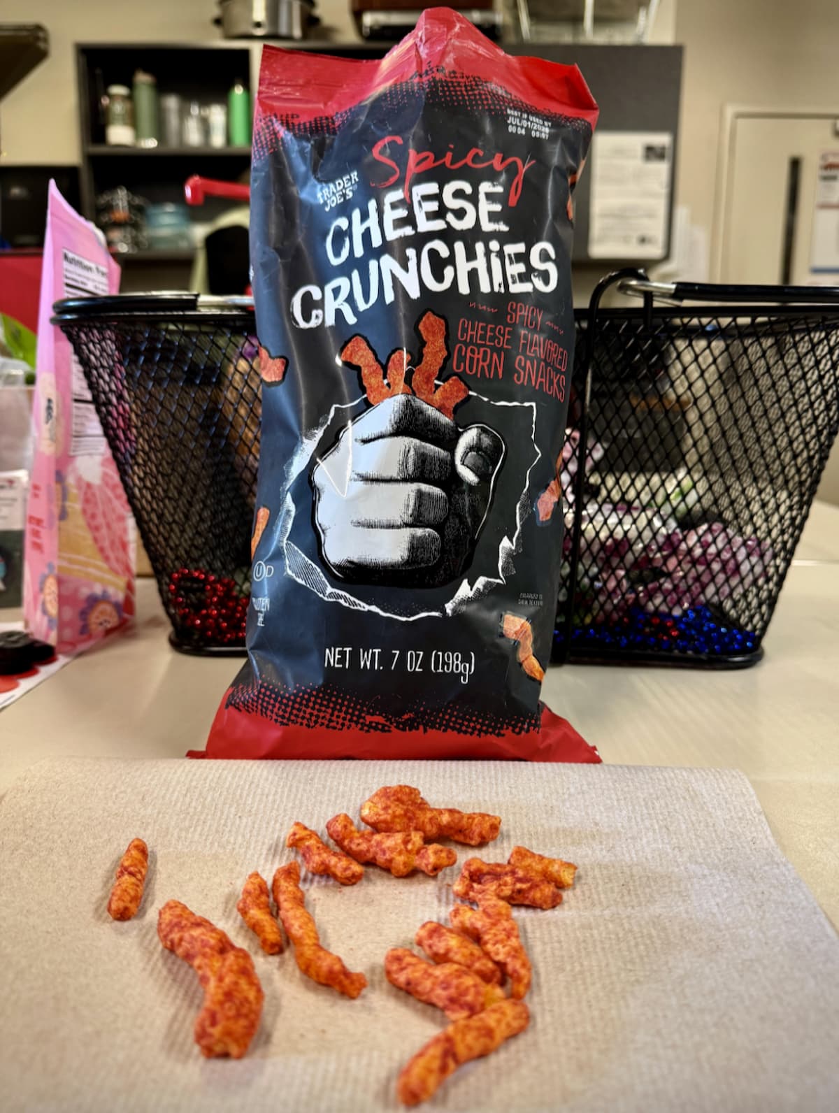 A bag of Spicy Cheese Crunchies sits on a table, partially open, with several crunchy cheese snacks scattered on a napkin in front. The background shows wire baskets and kitchen items.