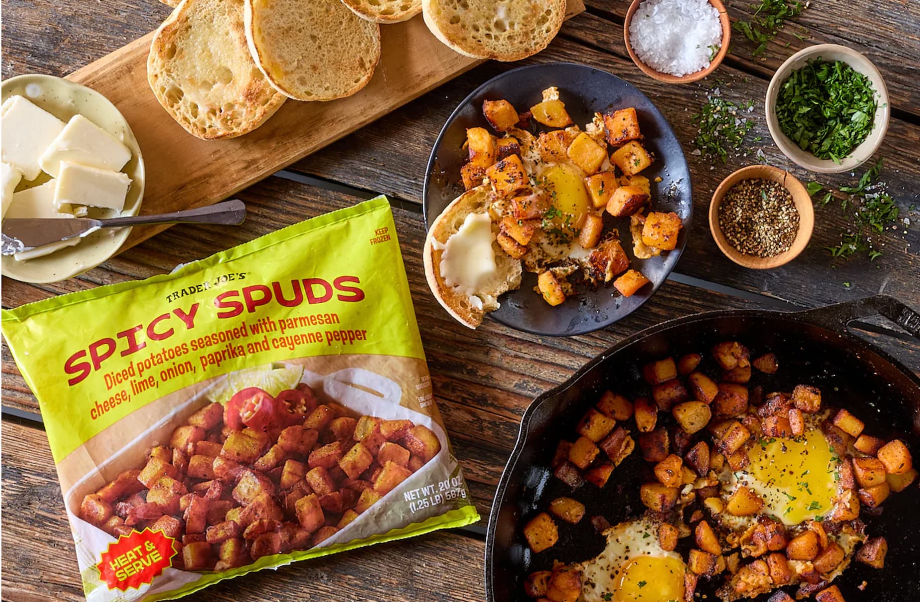 A bag of Trader Joe’s Spicy Spuds sits on a wooden table next to a skillet with cooked potatoes and eggs, butter, toast, fresh herbs, spices, and a small dish of coarse salt.