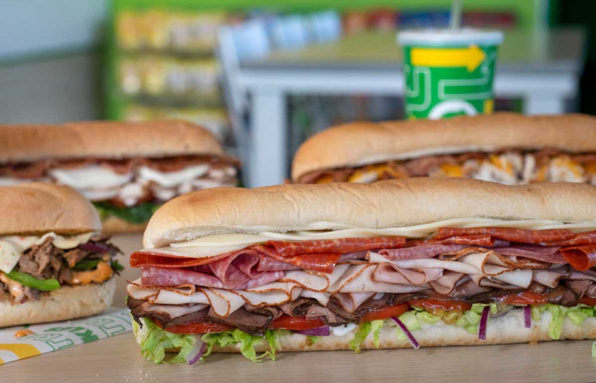 Several large sub sandwiches filled with meats, cheese, and vegetables are displayed on a table. In the background, a soft drink cup with a straw and blurred chairs and shelves are visible.