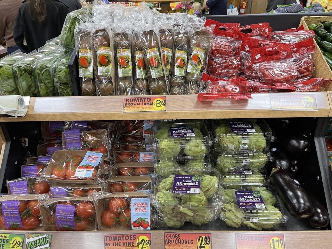 Supermarket produce shelf with Kumato brown tomatoes, grape tomatoes, cherry tomatoes, globe artichokes, eggplants, and green grapes in clear packaging, with colorful price tags below.