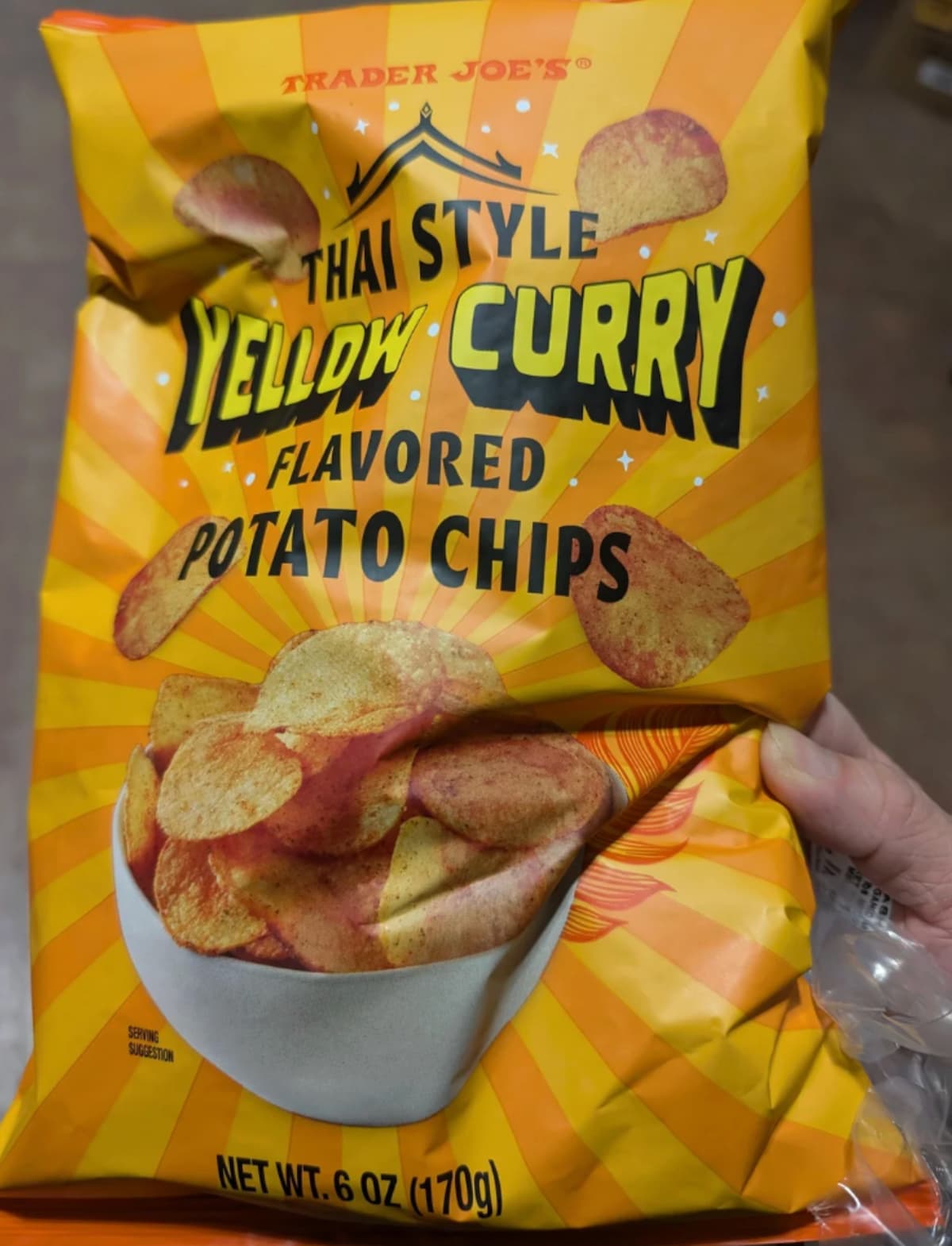 A hand holds a 6 oz (170g) bag of Trader Joe's Thai Style Yellow Curry Flavored Potato Chips, with chips pictured in a white bowl on an orange-yellow striped background.