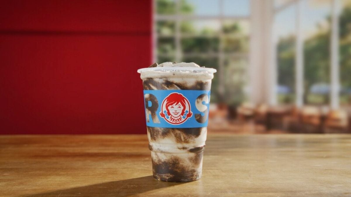 A new fast-food menu favorite: a Wendy's Frosty drink with chocolate and vanilla swirls in a clear cup featuring the Wendy's logo, placed on a wooden table with a blurred indoor background.