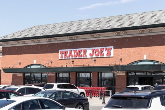 The image shows the exterior of a Trader Joe's store with a large sign above the entrance. Several parked cars and red shopping carts are visible in front of the building.