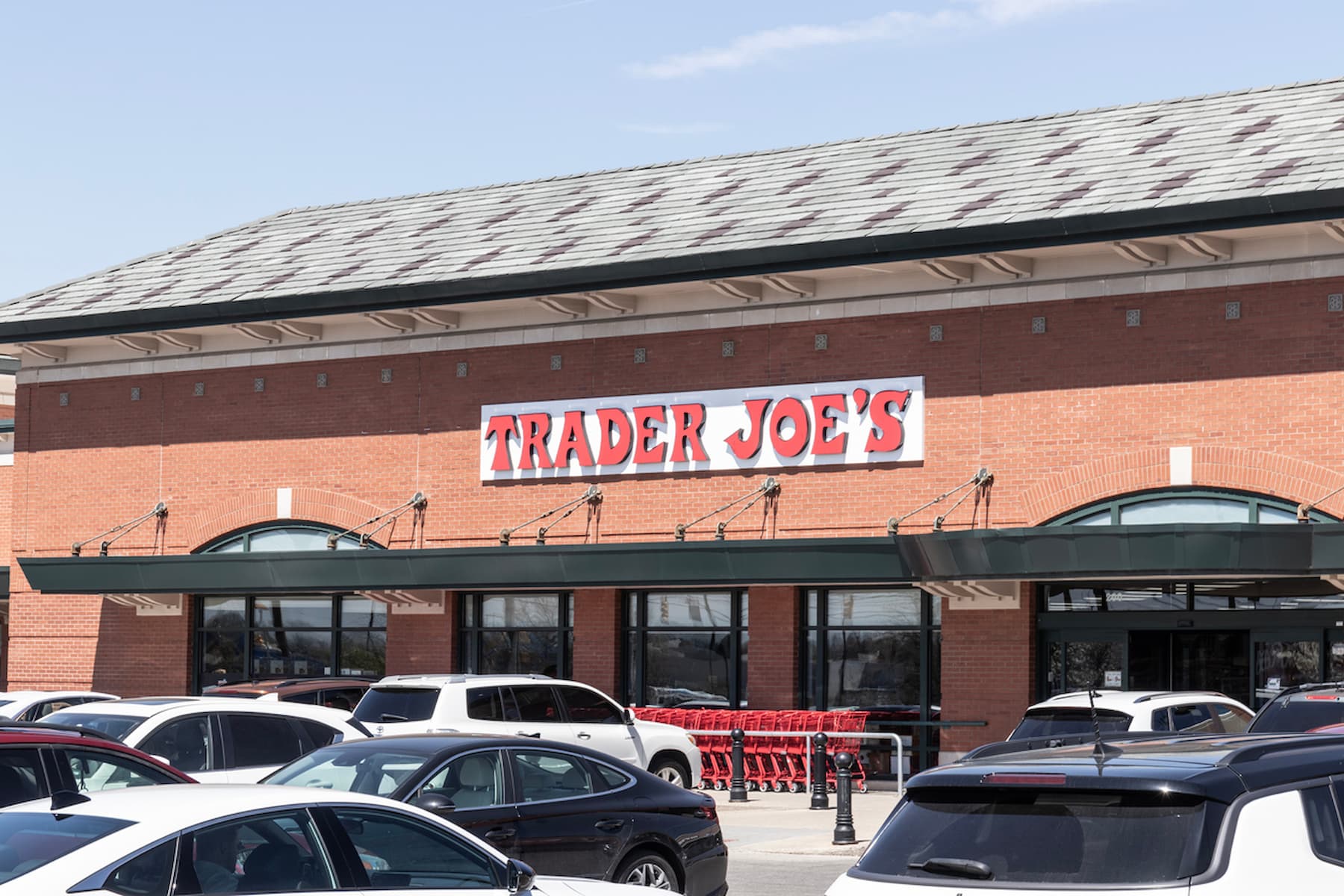 The image shows the exterior of a Trader Joe's store with a large sign above the entrance. Several parked cars and red shopping carts are visible in front of the building.