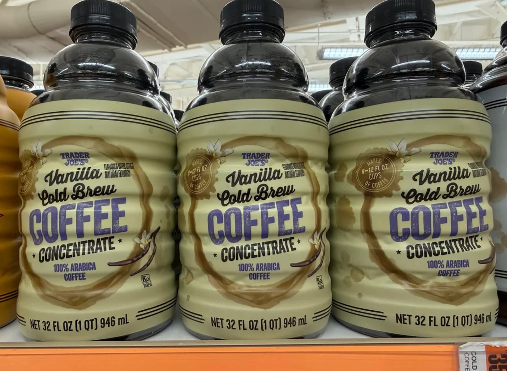 Three bottles of Trader Joe’s Vanilla Cold Brew Coffee Concentrate are lined up on a store shelf. Each bottle is labeled as 32 fl oz (946 ml) and features a beige and brown design with a coffee cup illustration.